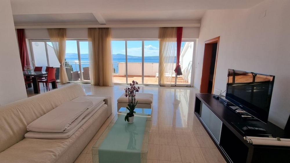 Pohľad na svetlý gauč, TV a more a pobrežie v pozadí.