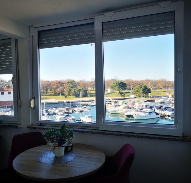 Útulný jedálenský kút s okrúhlym stolom a výhľadom na prístav cez veľké okno v chorvátskom apartmáne - Kúpte si obytný priestor v Chorvátsku.