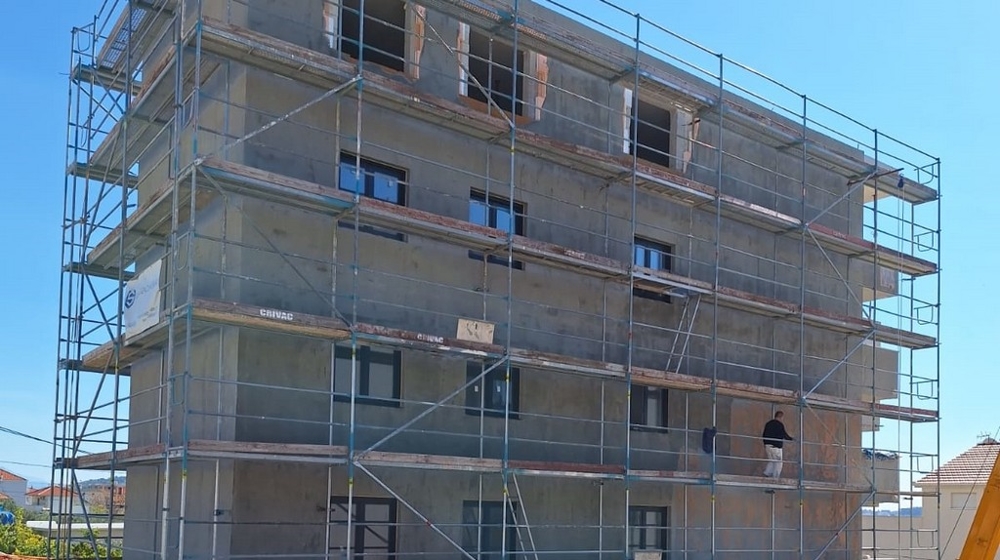 Bytový dom vo fáze výstavby s lešením, viditeľným plášťom a prácami na obvodovom murive, modrá obloha v pozadí.