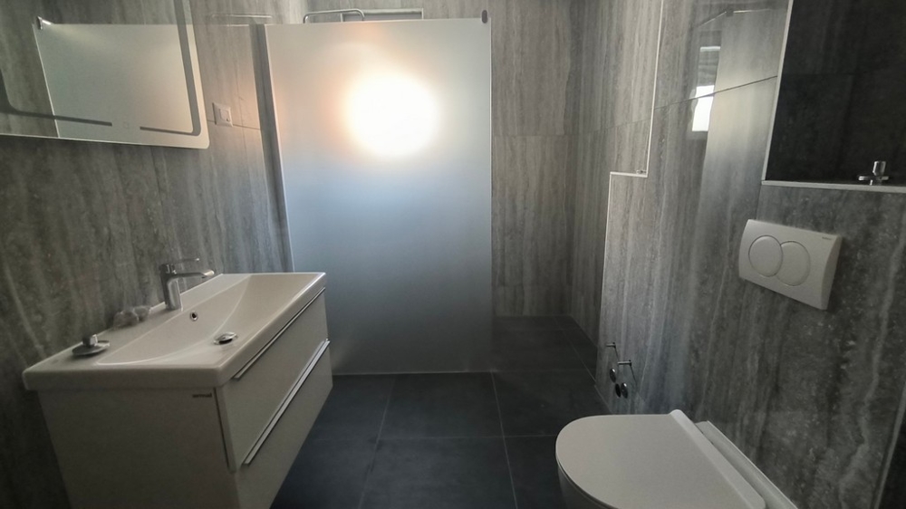 Moderne navrhnutá kúpeľňa so sprchovacím kútom, nehnuteľnosť v Chorvátsku v Šimuni, zdôraznená sivými dlaždicami a jasným svetlom.