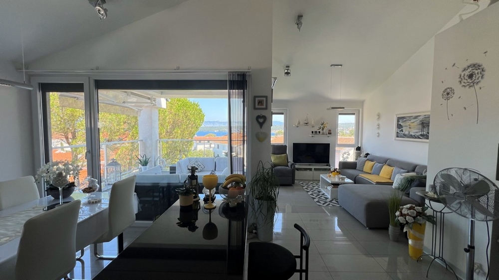 Svetlá obývacia izba so sivou pohovkou, jedálenským kútom a veľkým oknom na terasu s výhľadom na more.