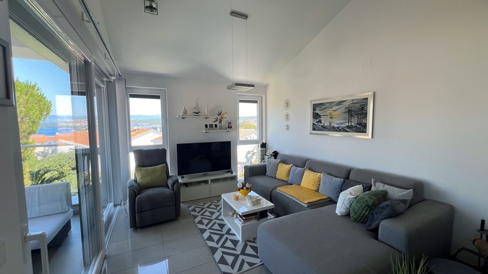 Moderná obývacia izba s veľkou TV, sivou pohovkou a výhľadom na more cez veľké okná.