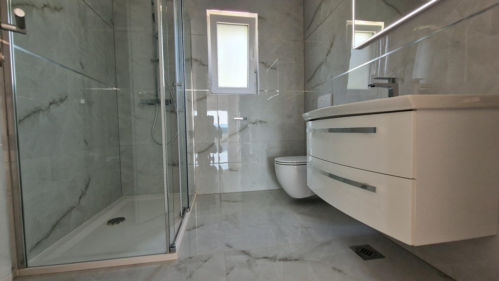 Moderná kúpeľňa so sprchovacím kútom, WC a umývadlom, svetlá dlažba a okná.