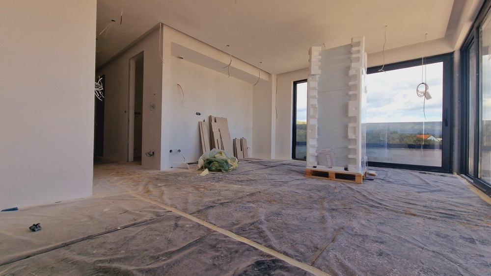 Obytná časť novostavby vo fáze dokončovania, s výhľadom na veľké presklené okná a okolitú prírodu.