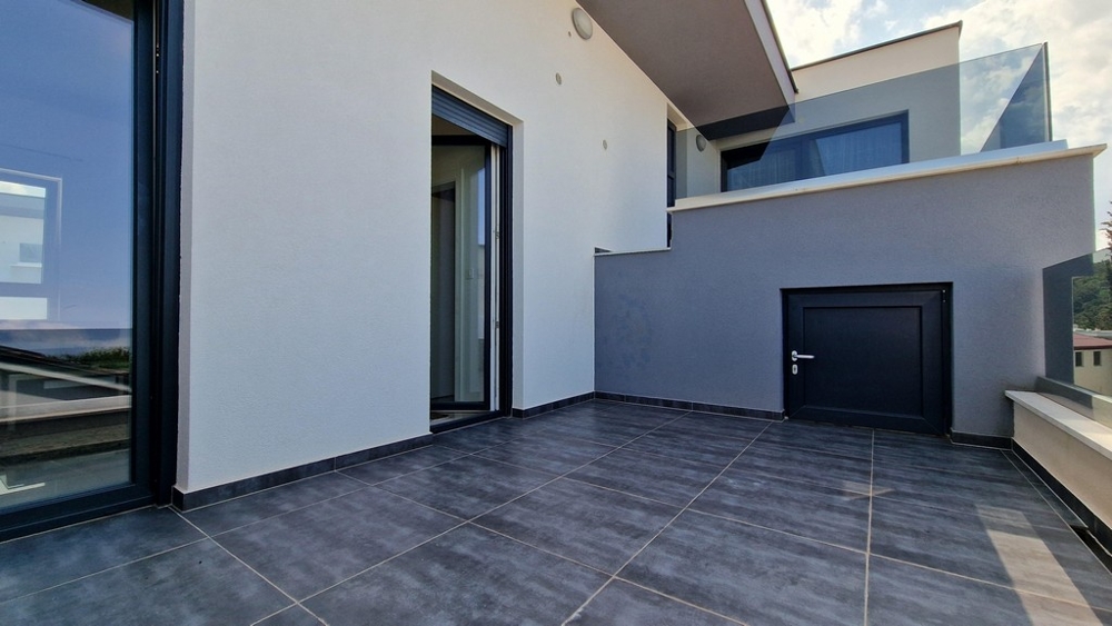 Moderná terasa so sivou dlažbou, skleneným zábradlím a vstupom do bytu.