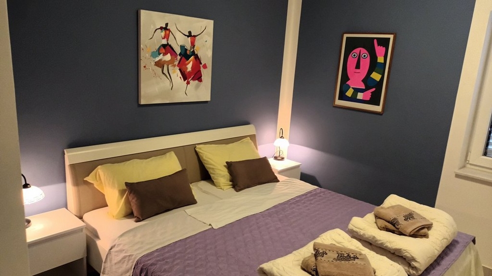 Spálňa s manželskou posteľou, fialovou perinou a dvoma obrázkami na stenách.