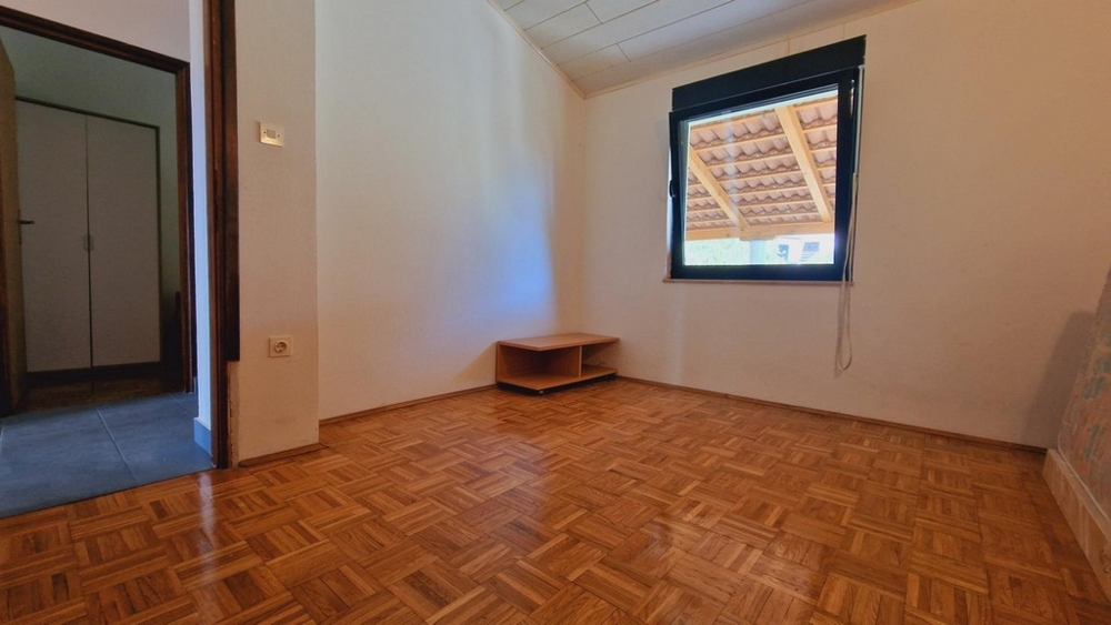 Prázdna spálňa s veľkým oknom, jednoduchým nábytkom a drevenou podlahou.