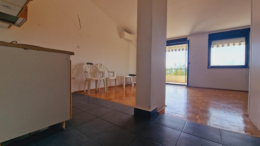 Obývacia izba s otvoreným prechodom do kuchyne, jednoduchý biely nábytok a dlažba.