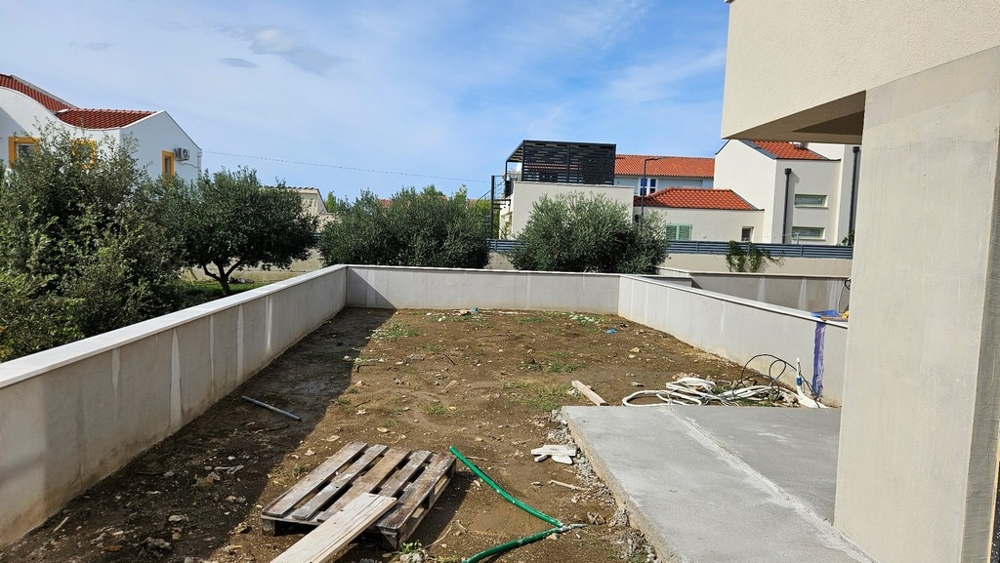 Vonkajší priestor s nespevnenou zemou, nízkym múrikom a niekoľkými stromami v pozadí.