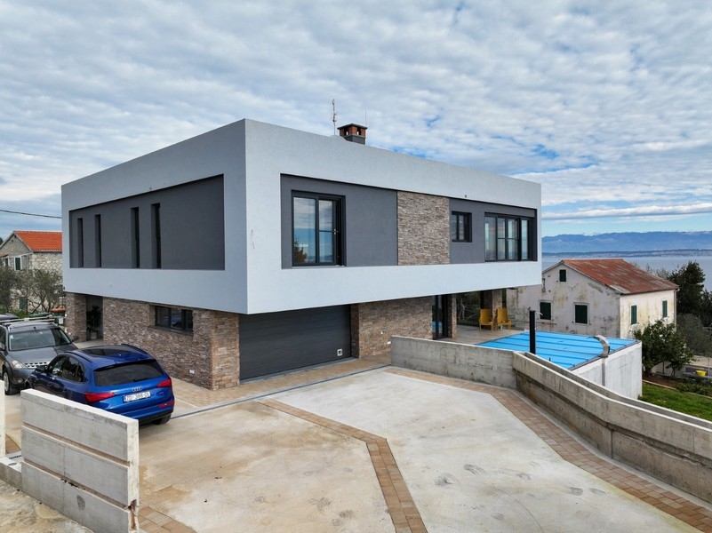 Pohľad spredu na modernú vilu s garážou na predaj na Ugljan v Chorvátsku, obklopenú dláždenou príjazdovou cestou a čiastočným výhľadom na more
