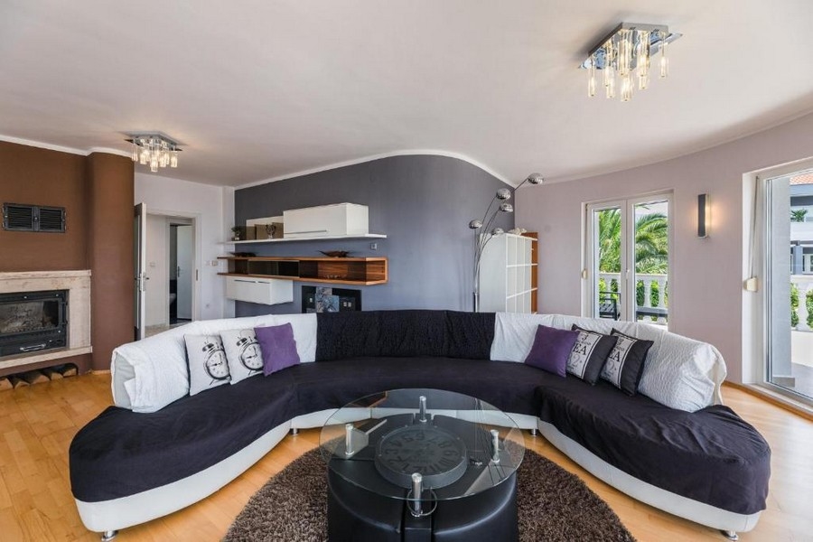 Elegantná obývacia izba v chorvátskom apartmánovom dome s výhľadom na more, s veľkou zakrivenou pohovkou a okrúhlym skleneným stolom na hnedom koberci, krbom a otvoreným balkónom.