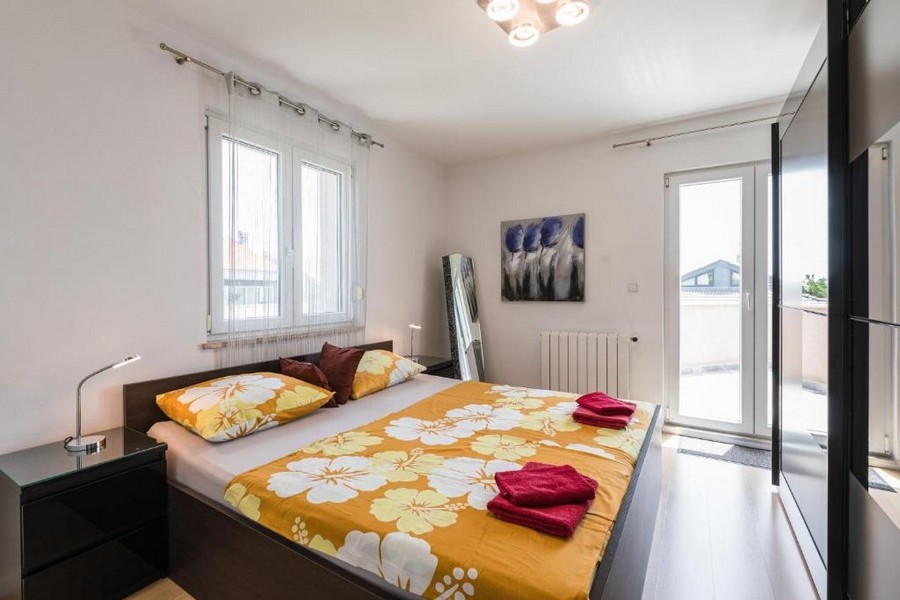 Moderná spálňa v chorvátskom apartmánovom dome s výhľadom na more, s manželskou posteľou so žltou posteľnou bielizňou, nočnými stolíkmi a umením na stene, prístupom na terasu