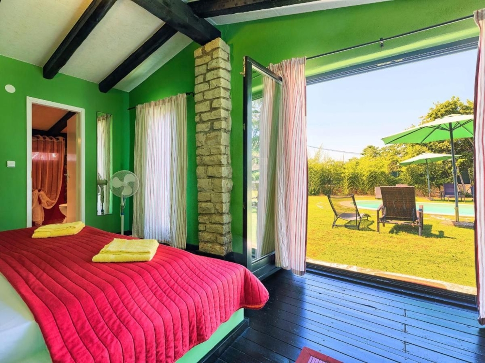 Svetlá spálňa s výhľadom do záhrady, súčasť nehnuteľnosti v Chorvátsku, so zelenými stenami, červenou prikrývkou a prístupom na terasu s ležadlami a slnečníkom