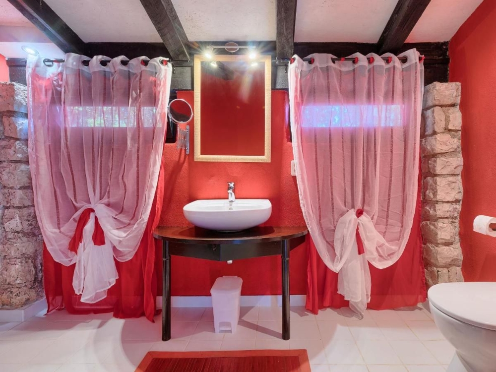Kúpeľňa nehnuteľnosti v Chorvátsku s červenými stenami, bielou inštalatérstvom, umývadlom na rustikálnom stole a priehľadnými červenými závesmi na oknách