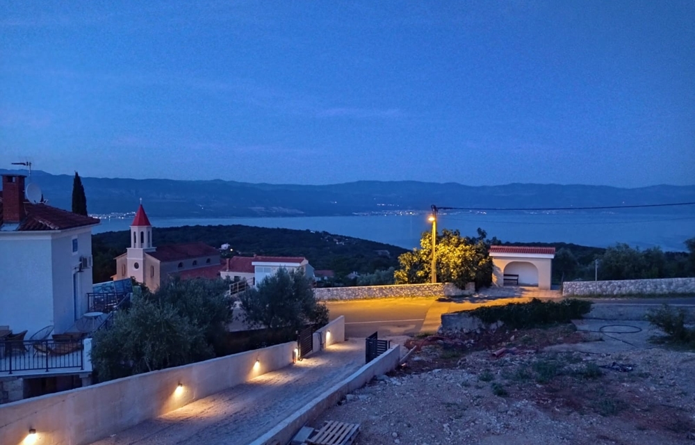 Pohľad z nehnuteľnosti v Chorvátsku na more v noci, bazén v popredí a osvetlenú dedinu v pozadí.