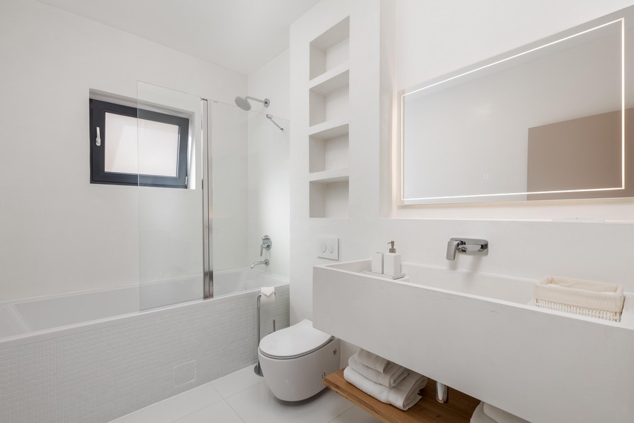 Jednoduchá kúpeľňa s bielou podlhovastou doskou, vstavanými policami, vaňou so sprchovacím nástavcom a WC. Niektoré steny majú biele dlaždice a veľké zrkadlo.