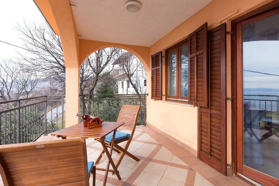 Krytý balkón s drevenými stoličkami a stolom s výhľadom na zelené okolie a more v diaľke.