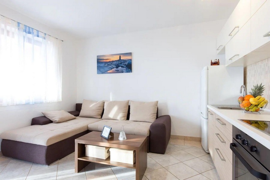 Útulná obývačka s rohovou pohovkou, konferenčným stolíkom a výhľadom do kuchyne, zariadená v jemných farbách a s obrazom mora.