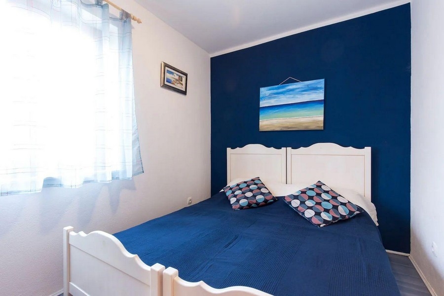 Spálňa s manželskou posteľou, modrou akcentovanou stenou a námornou dekoráciou, osvetlená prirodzeným svetlom z okna.