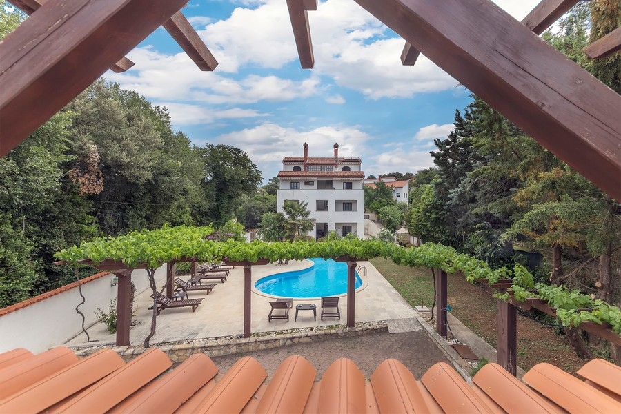 Pohľad z pergoly na bazénovú časť a biely bytový dom obklopený bujnou vegetáciou