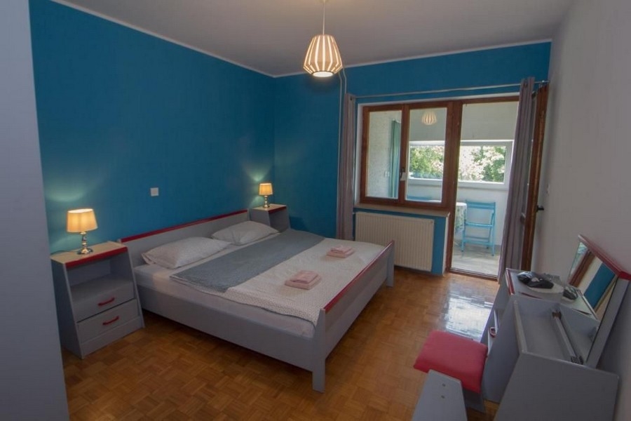 Spálňa s modrými stenami, veľká manželská posteľ, dva nočné stolíky s lampami, okno a dvere vedúce na balkón.