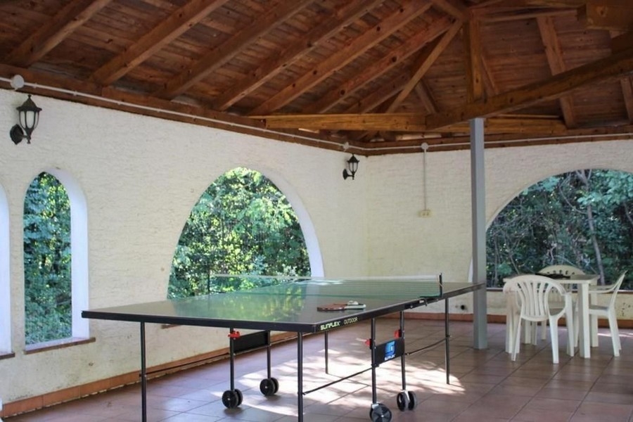 Krytý vonkajší priestor so stolným tenisom uprostred, bielymi plastovými stoličkami a drevenou strechou. Steny majú klenuté okenné otvory s výhľadom do zelene.