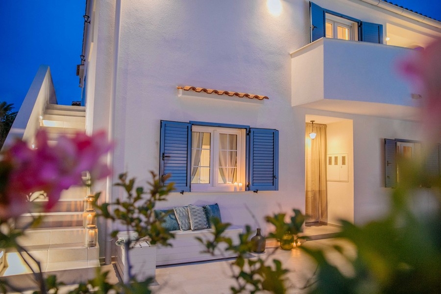 Pohľad spredu na stredomorský dom s modrými okenicami a vonkajším posedením za súmraku, osvetlený jemným svetlom