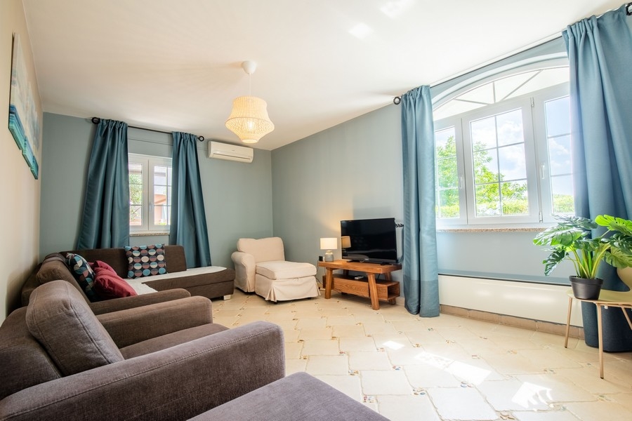 Útulná obývacia izba s niekoľkými pohovkami, TV a veľkými oknami s modrými závesmi, ktoré prepúšťajú prirodzené svetlo