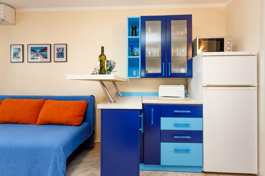 Kuchyňa s modrými skrinkami, bielou chladničkou a malým kuchynským riadom