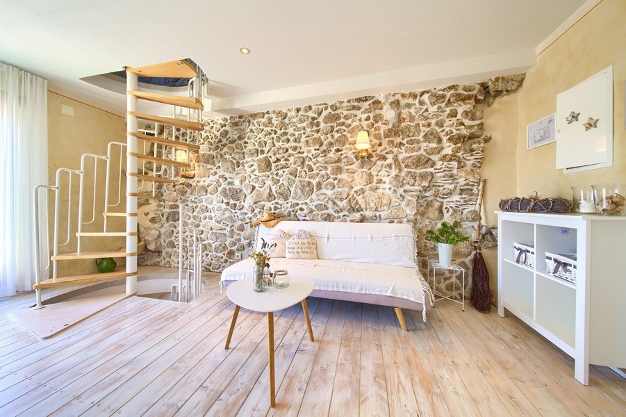 Útulná obývacia izba s odhalenými kamennými stenami, pohovkou a točitým schodiskom vedúcim na poschodie.