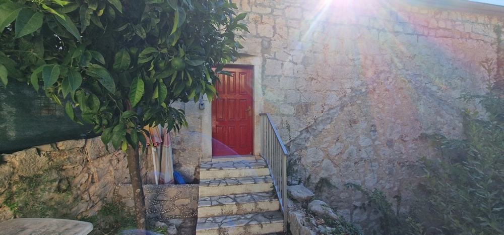 Malý vonkajší priestor s kamenným schodiskom vedúcim k červeným dreveným dverám. Stena domu je z prírodného kameňa a na oblohe svieti slnko.