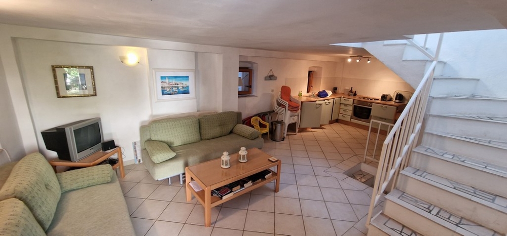 Útulný, otvorený obývací priestor so zelenými pohovkami, konferenčným stolíkom, TV a integrovanou kuchyňou v zadnej časti. Svetlé dlaždice pokrývajú podlahu.