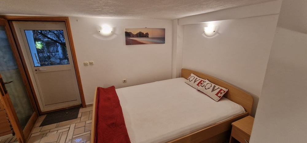 Spálňa s manželskou posteľou zdobená bielymi a červenými prvkami. Na stene je obraz pláže a von vedú dvere.